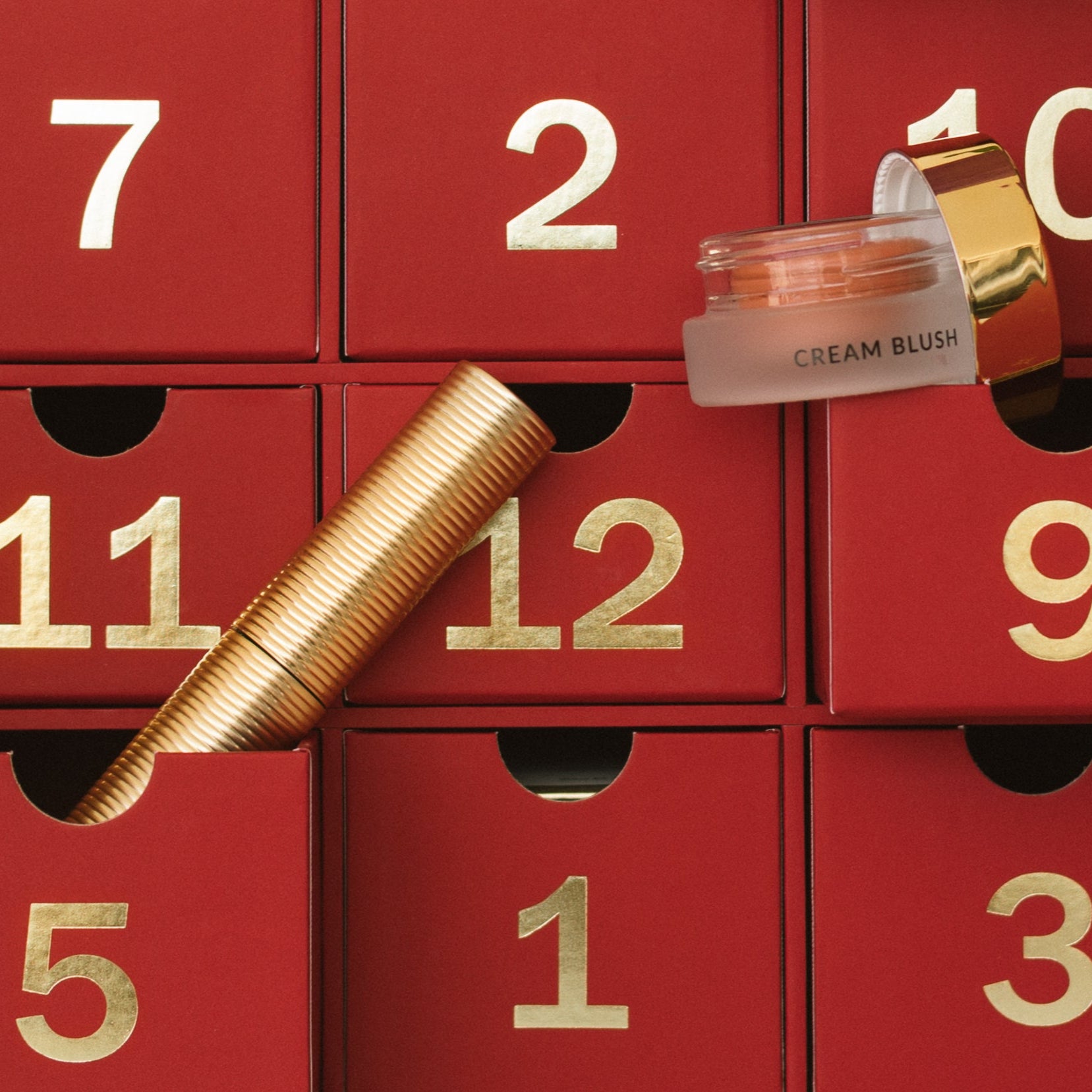 Red advent calendar with gold numbered drawers, featuring a cream blush and gold tube mascara.
