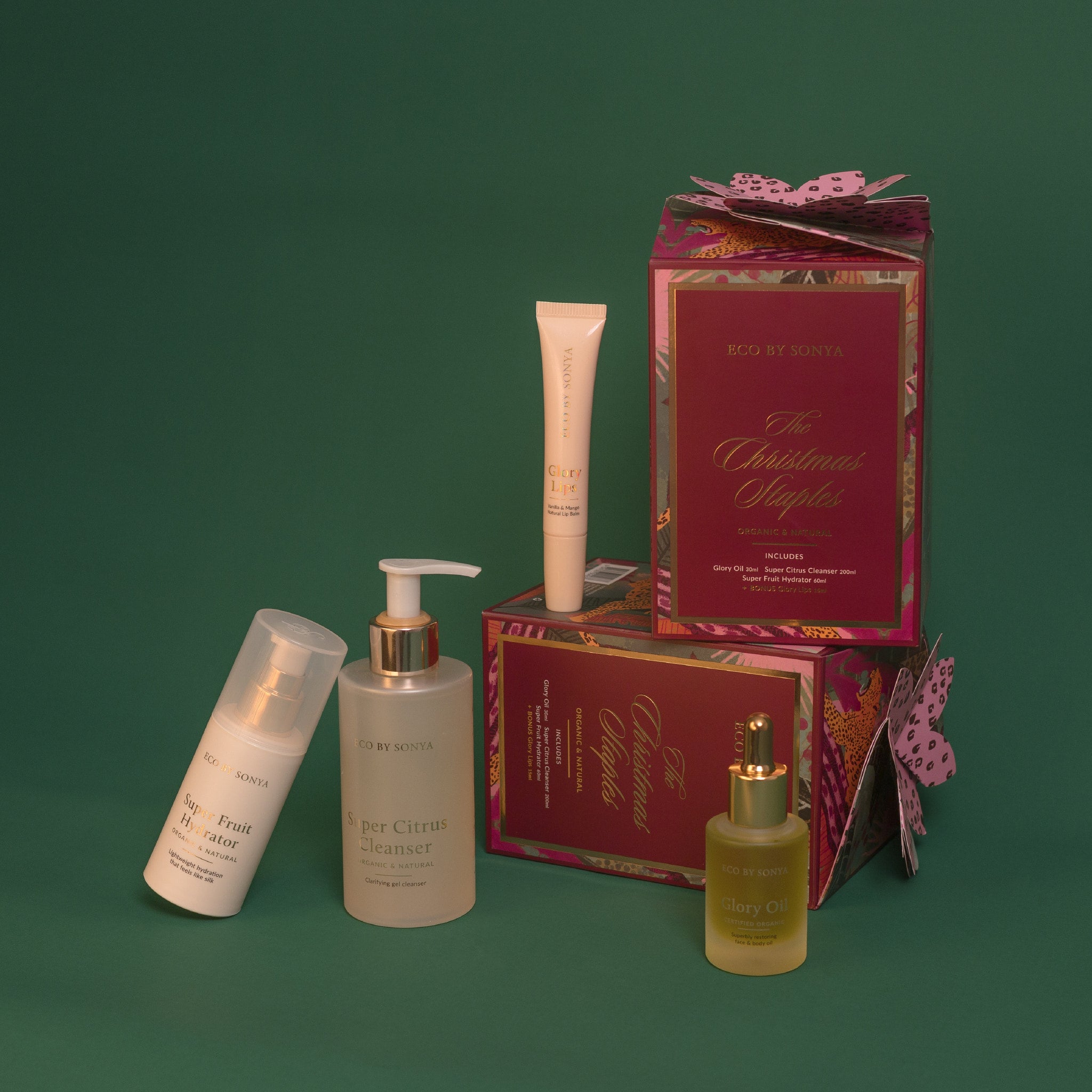 co By Sonya The Christmas Staples Gift Set featuring Super Citrus Cleanser, Super Fruit Hydrator, Glory Oil and bonus Glory Lips, arranged with decorative red and gold gift boxes on a dark green background.
