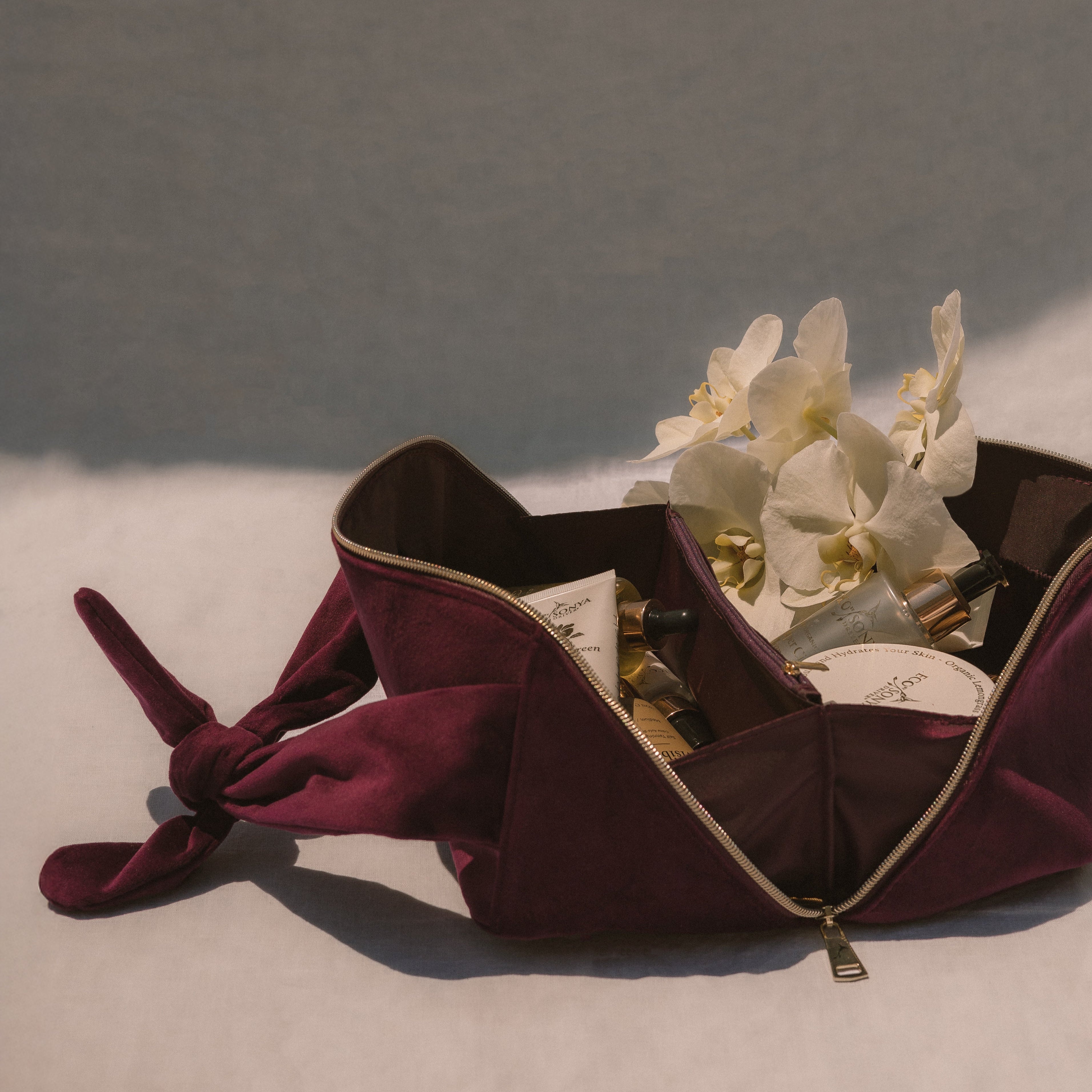 Burgundy travel cosmetic bag open with products inside, white orchids