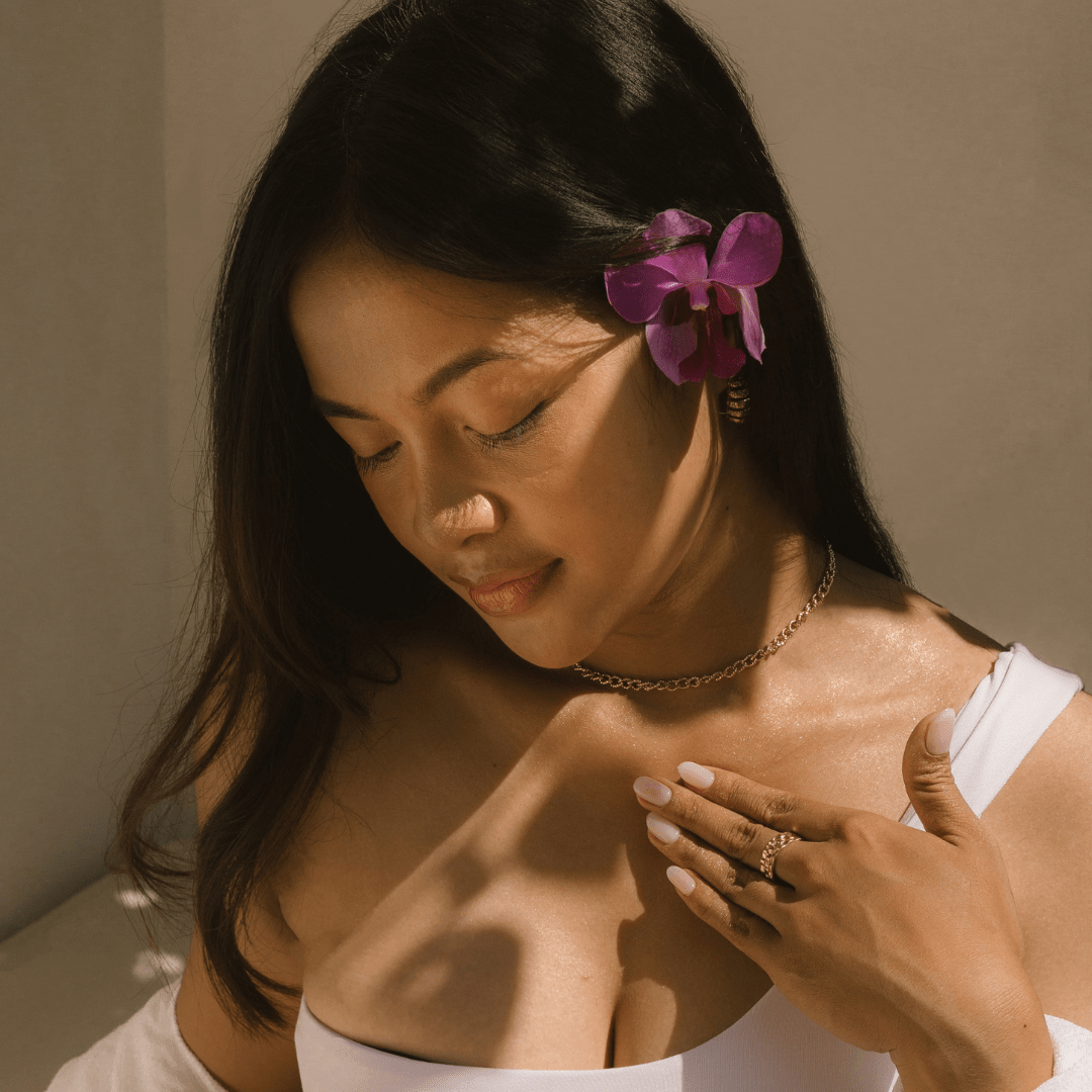 Woman with a purple flower in her hair, wearing a white top, applying Golden Illuminiser highlighting drops to her decolletage.