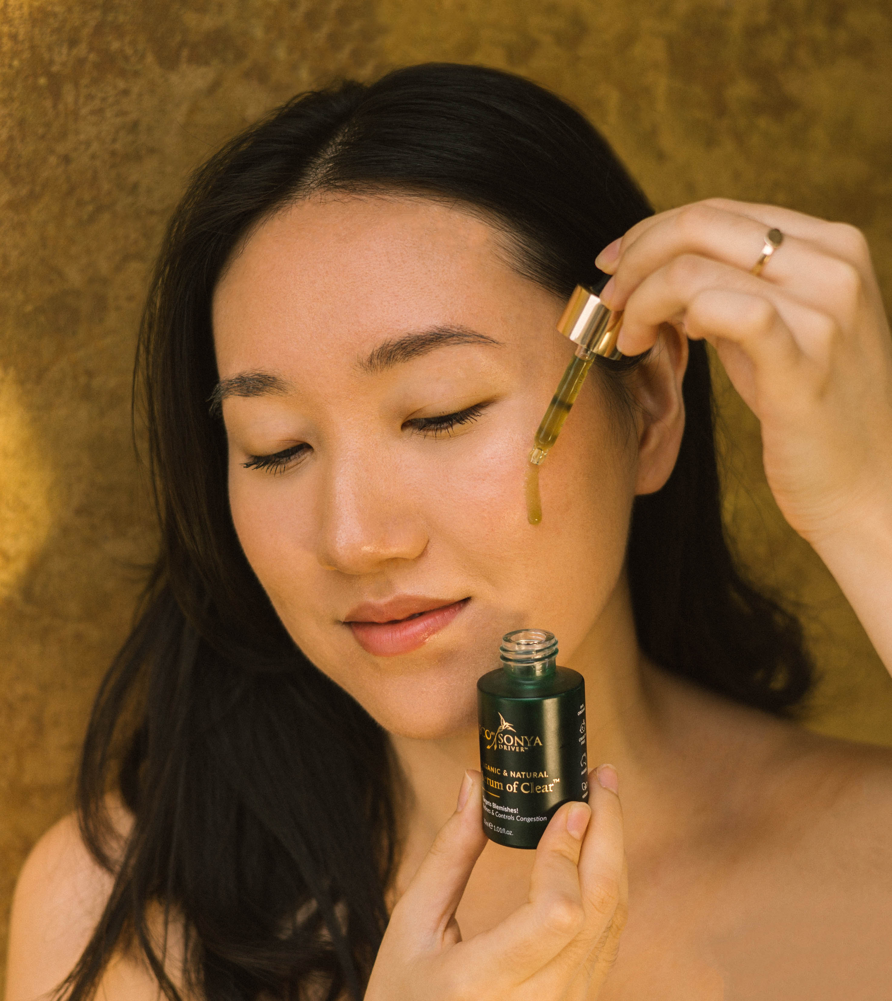 Woman applying a dropper of Serum of Clear to her face with a gold background