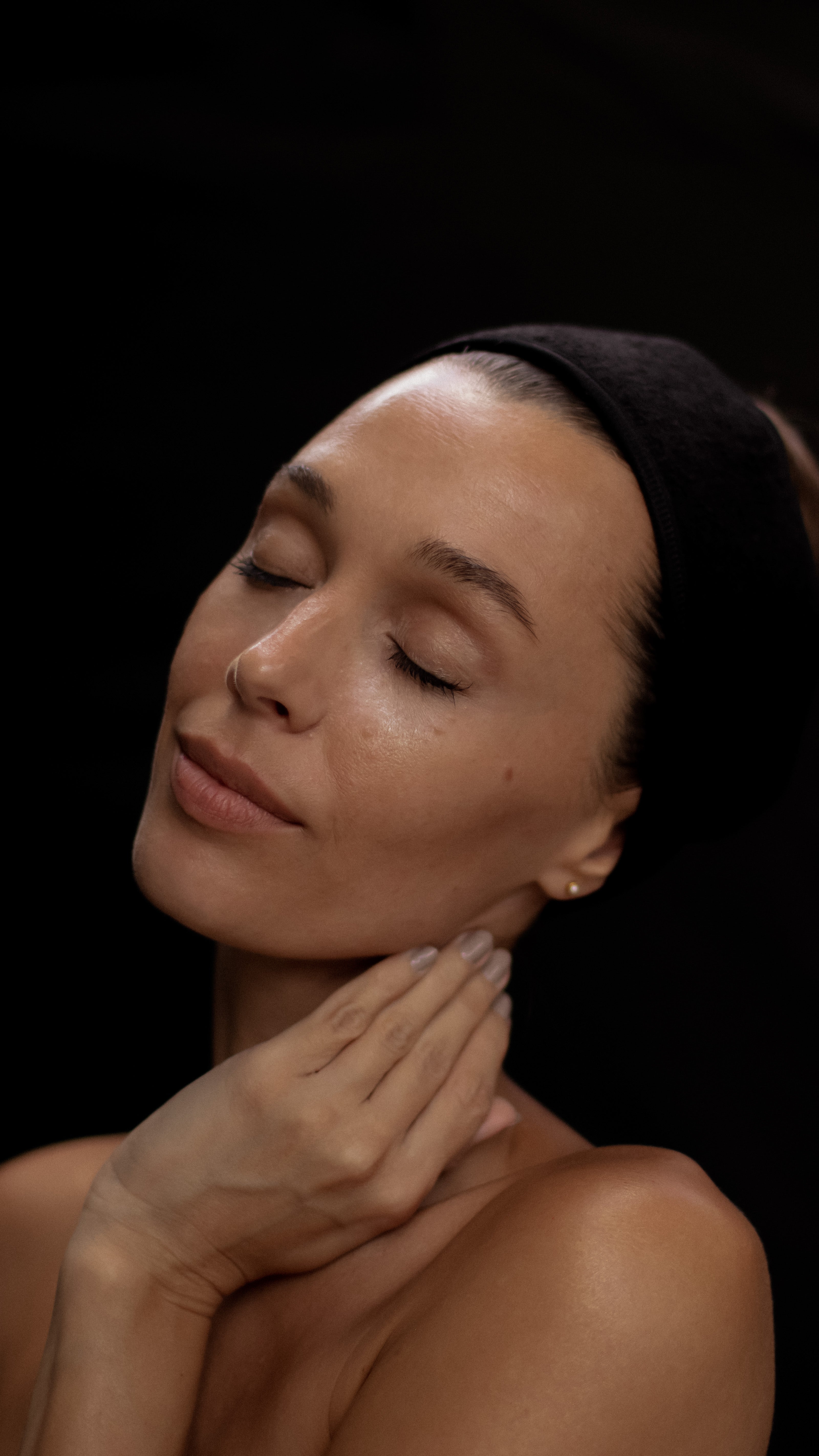 Woman with closed eyes touching her neck against a black background