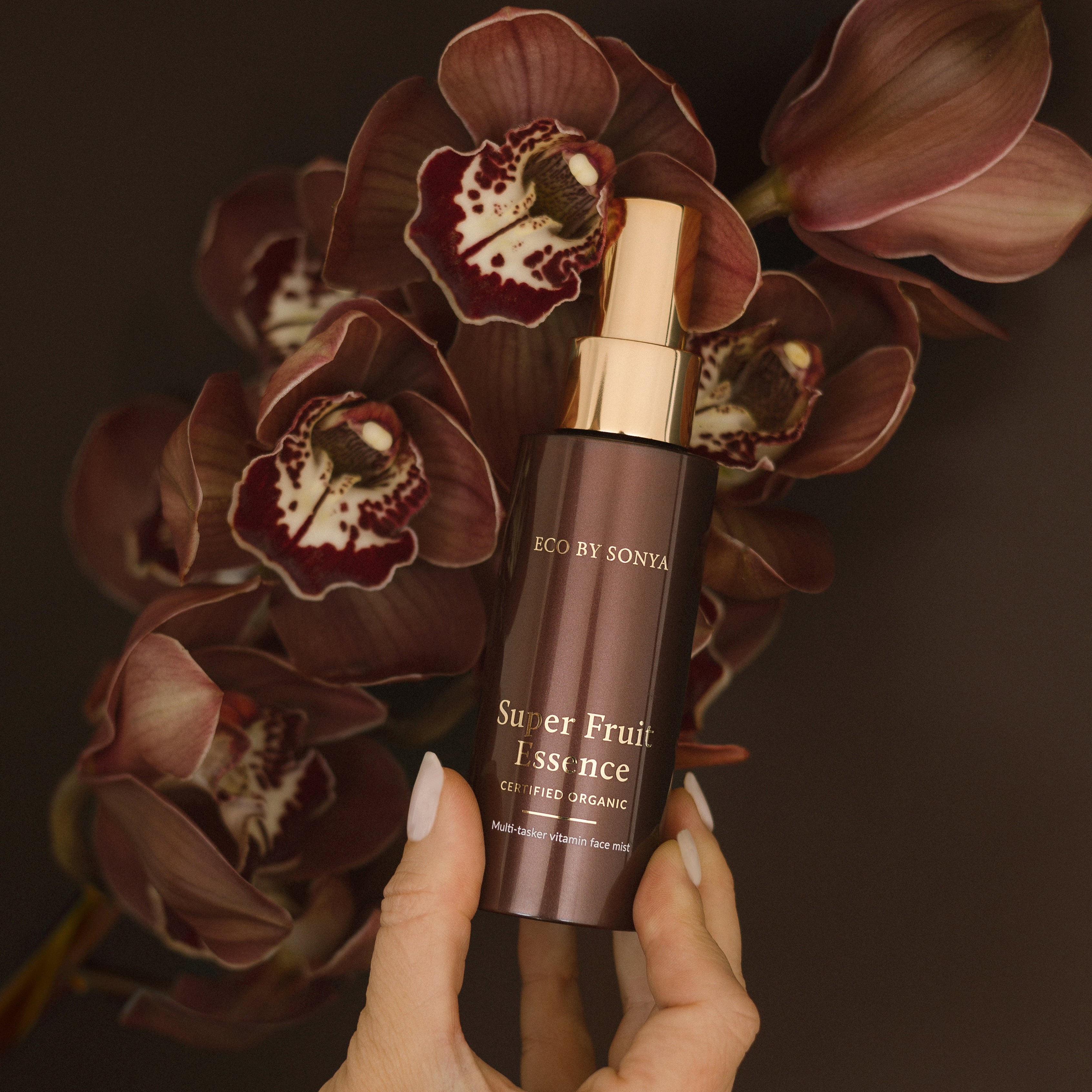 Hand holding a bottle of Eco By Sonya Super Fruit Essence' against a dark background with flowers