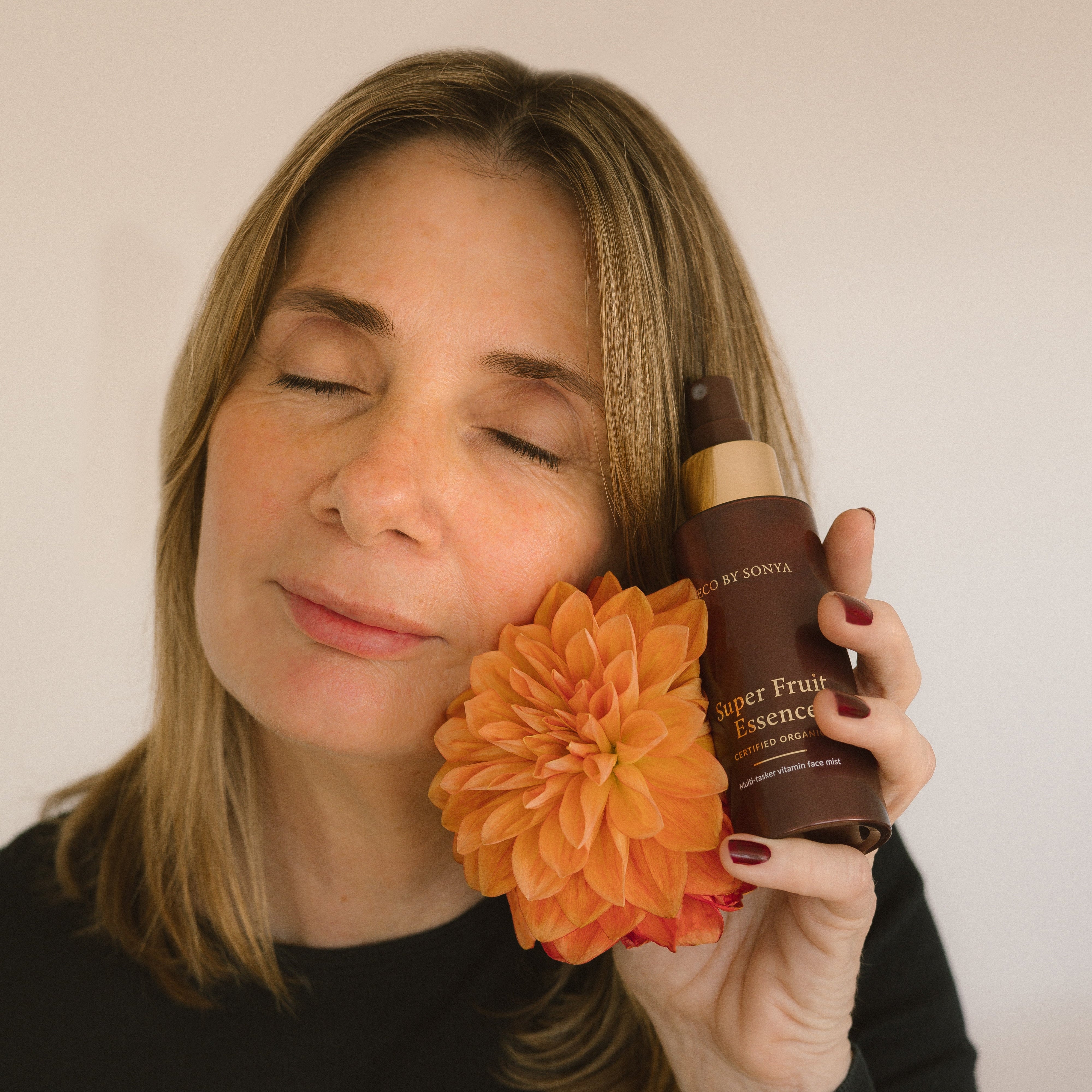 Woman holding an orange flower and a bottle of Eco By Sonya Super Fruit Essence against a plain background