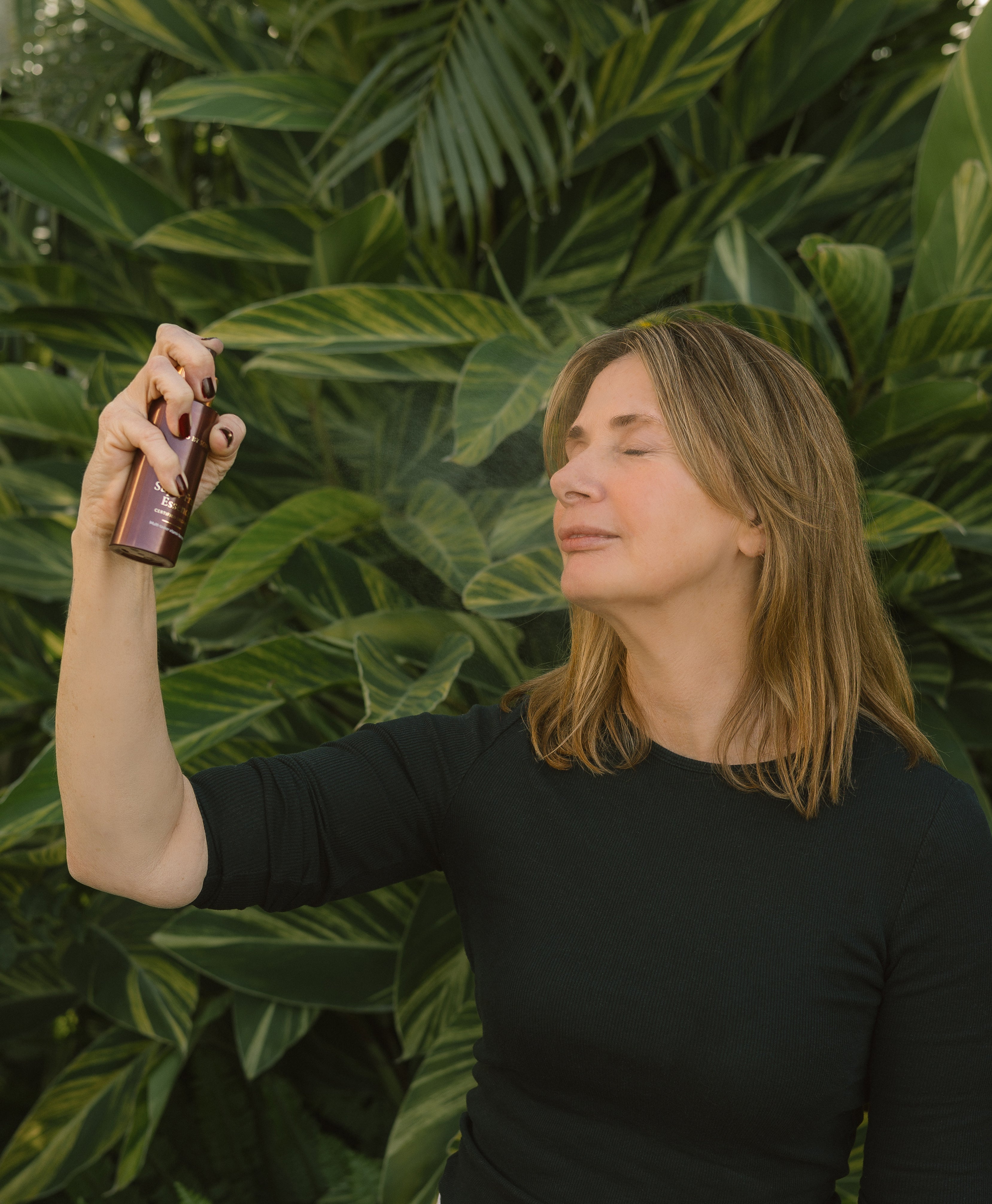 Woman spraying Super Fruit Essence onto her face, against a green leafy background